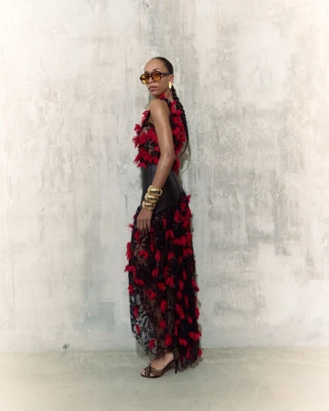 Elegant woman poses in a black and red feather dress against a textured wall. She wears sunglasses and gold bracelets.