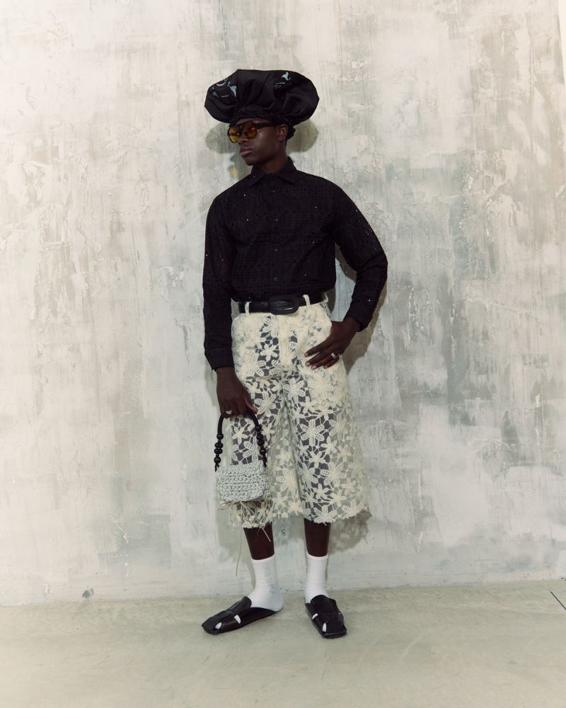 Man in a black shirt, lace shorts, oversized hat, sunglasses, socks, and sandals posing in a photo studio against a textured wall.