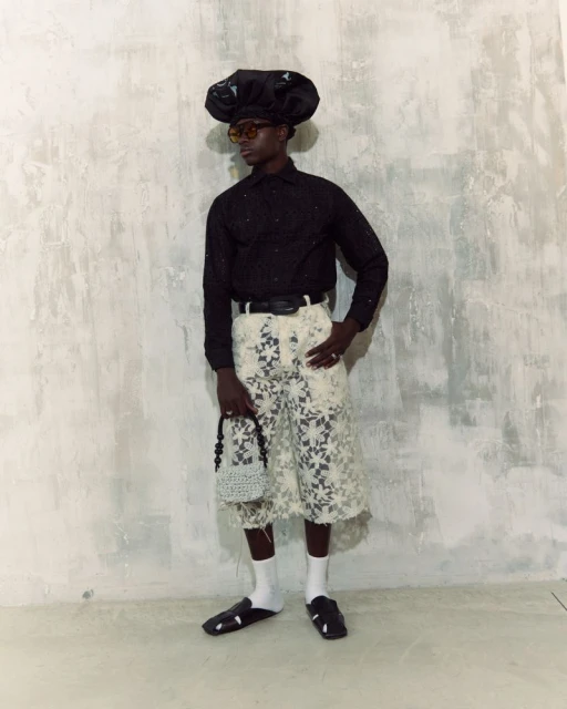 Man in a black shirt, lace shorts, oversized hat, sunglasses, socks, and sandals posing in a photo studio against a textured wall.