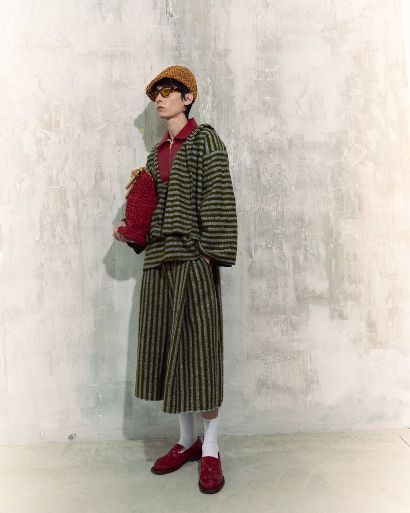 Stylish young adult in a striped olive-green co-ord set, accessorized with a hat, bag, glasses, socks, and loafers against a concrete wall.