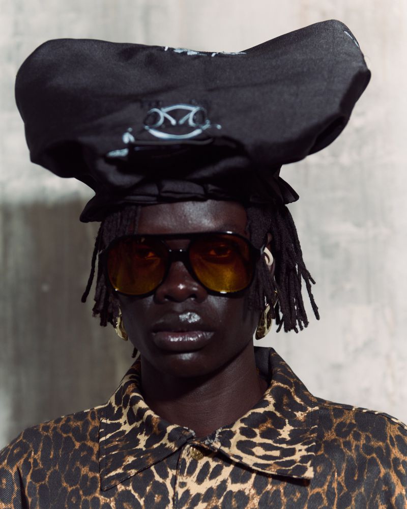 Close-up of a dark-skinned adult with dreadlocks wearing sunglasses, a black hat, and a leopard-print jacket against a gray background.