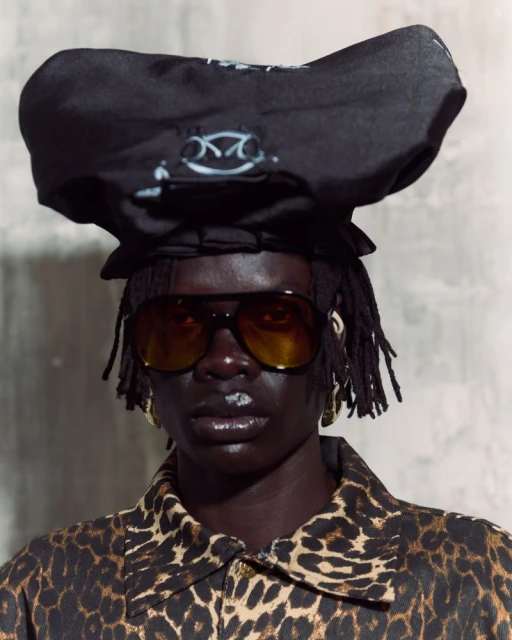 Close-up of a dark-skinned adult with dreadlocks wearing sunglasses, a black hat, and a leopard-print jacket against a gray background.