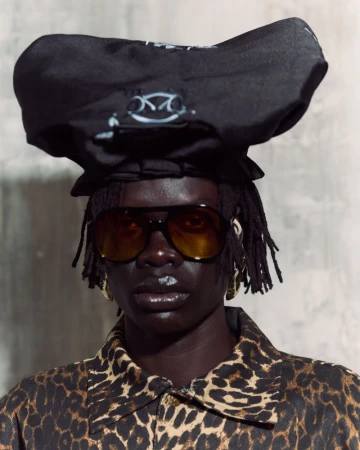 Close-up of a dark-skinned adult with dreadlocks wearing sunglasses, a black hat, and a leopard-print jacket against a gray background.