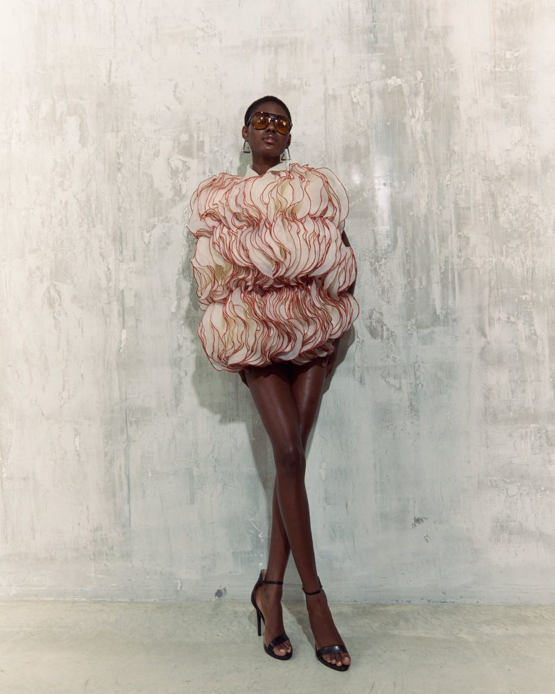 Woman in avant-garde ruffled mini dress and heels poses against a textured wall in a fashion shot with studio lighting.