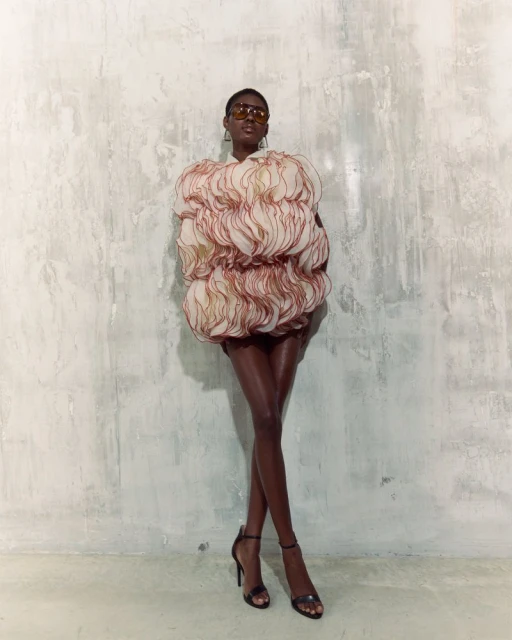 Woman in avant-garde ruffled mini dress and heels poses against a textured wall in a fashion shot with studio lighting.