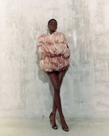 Woman in avant-garde ruffled mini dress and heels poses against a textured wall in a fashion shot with studio lighting.
