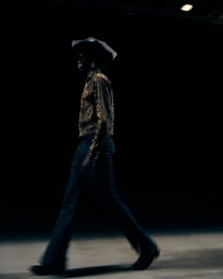 Model wearing a patterned shirt and dark trousers walking in a dimly lit backstage setting.