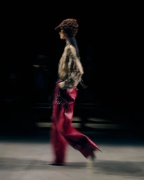 Model walking in profile wearing textured brown hat, patterned top, and red trousers against a dark background.
