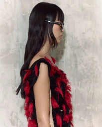 Profile view of a model wearing a black dress with red fringe accents and glasses against a textured concrete wall.