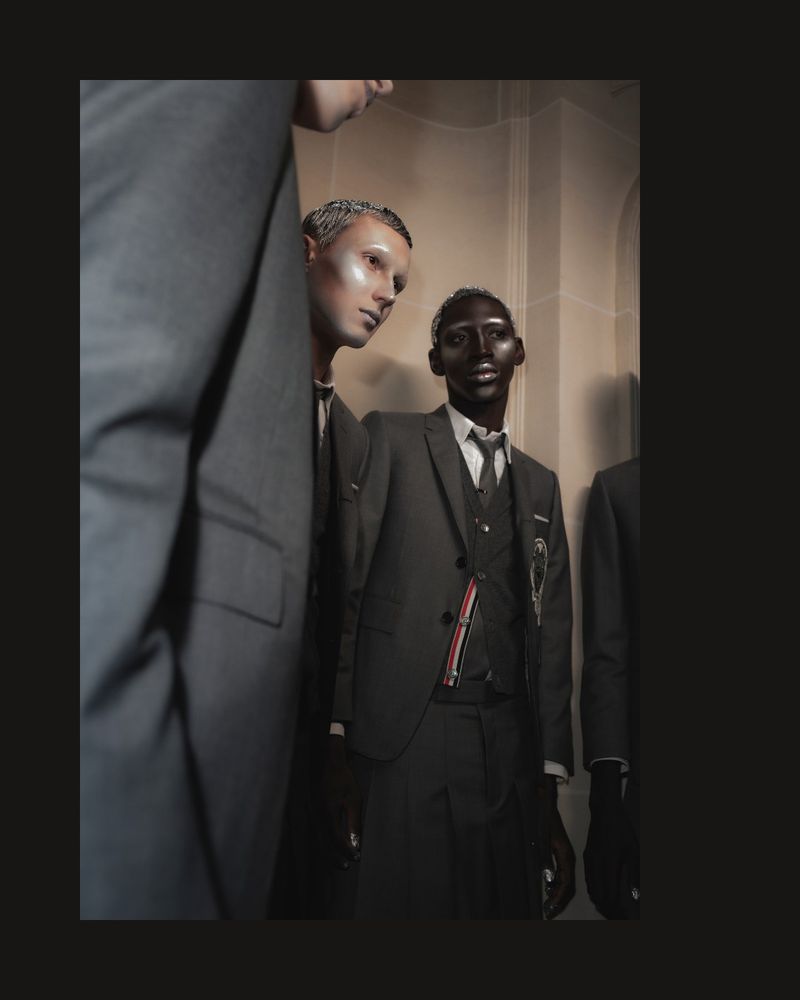 Two male models wearing dark suits and ties, posing in a dimly lit indoor setting, showcasing fashion and style with serious expressions.