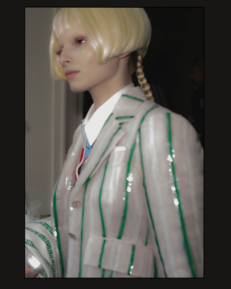 Fashion model wearing a white and green striped jacket and blonde braided wig in a photo studio with a dark background.