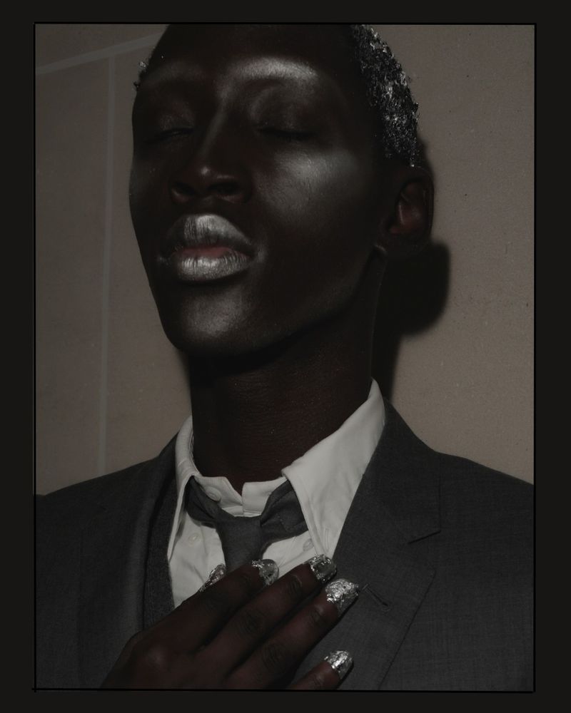 Close-up portrait of a person with dark skin, wearing a grey suit, white shirt, and tie, their face and hand decorated with shimmering silver.
