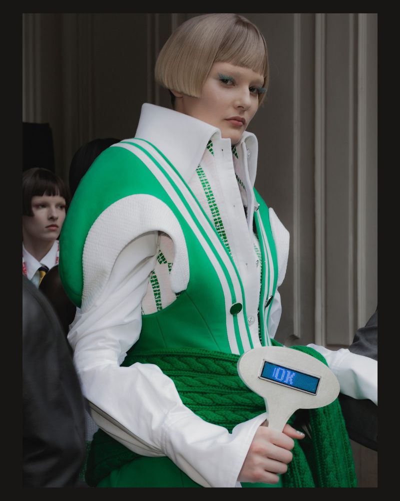 Fashion shoot featuring a young adult model wearing a green and white jacket, with bob hairstyle, holding an illuminated 'OK' sign.