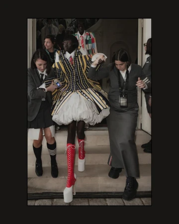 Runway model with dark skin wearing a yellow and black striped dress with red and white heeled boots, flanked by two attendants.