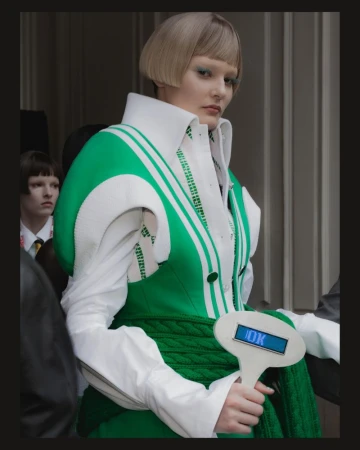 Model in green and white structured coat holding a paddle with a digital screen backstage at Thom Browne