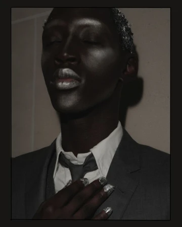 Portrait of a black male model with metallic makeup, silver hair, and a grey suit backstage at Thom Browne SS26.