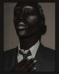 Portrait of a black male model with metallic makeup, silver hair, and a grey suit backstage at Thom Browne SS26.