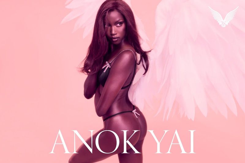Anok Yai poses in lingerie with angel wings against a light pink background in a beauty and fashion editorial portrait.