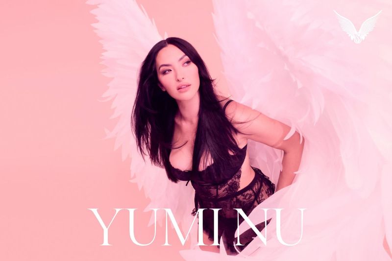 Portrait of a beautiful woman in black lingerie and large white feather wings posing against a soft pink backdrop in a photo studio.