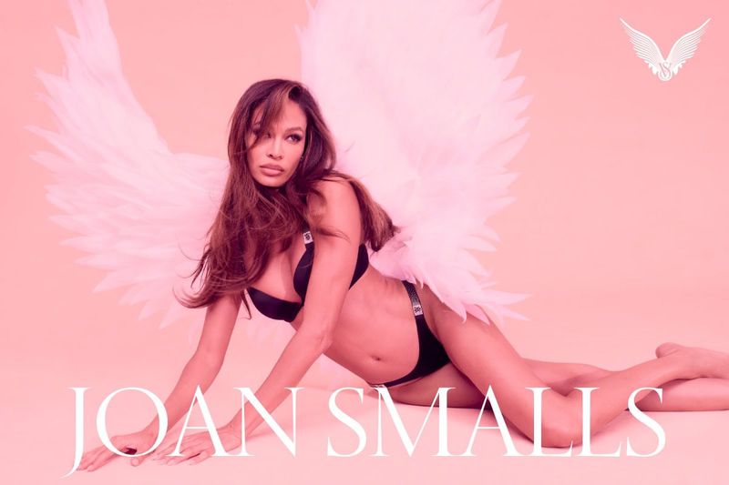 Joan Smalls poses in lingerie and angel wings against a peach background in a studio fashion shot with dramatic lighting.