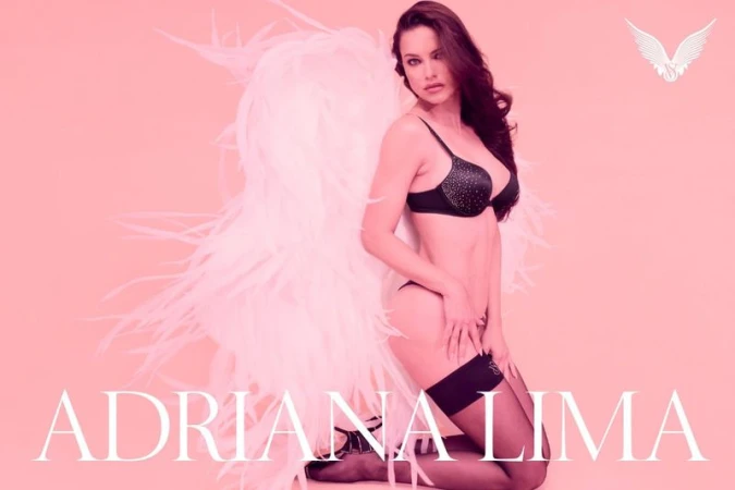 Adriana Lima wearing black lingerie with large white feathered angel wings against a pink background