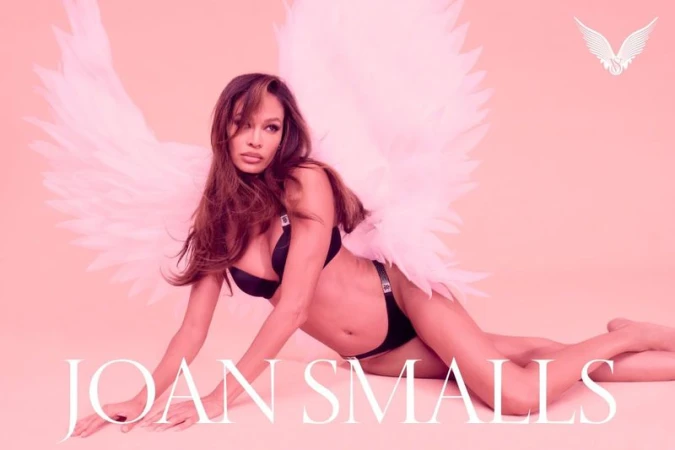 Model Joan Smalls wearing black lingerie and large white angel wings against a soft pink studio backdrop.