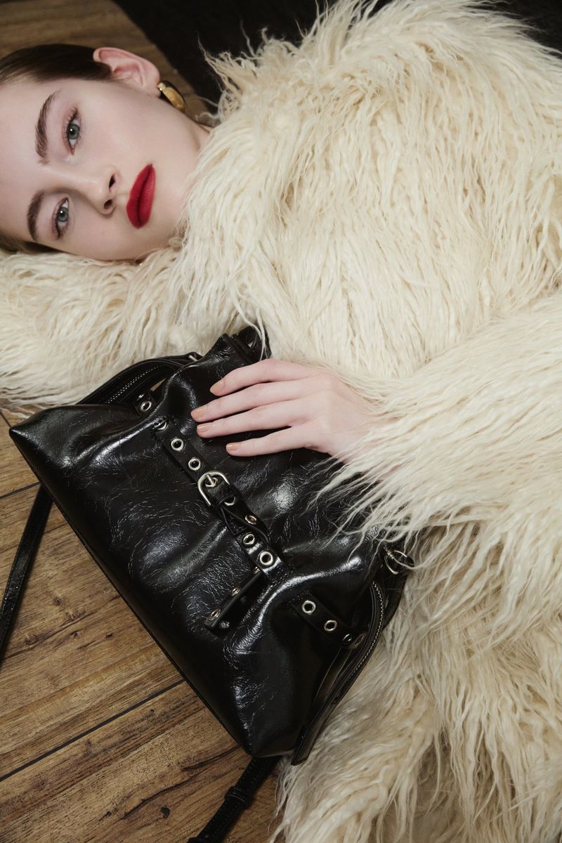 A fair-skinned model with dark red lipstick poses with a black purse against a cream-colored faux fur coat and wood flooring backdrop.