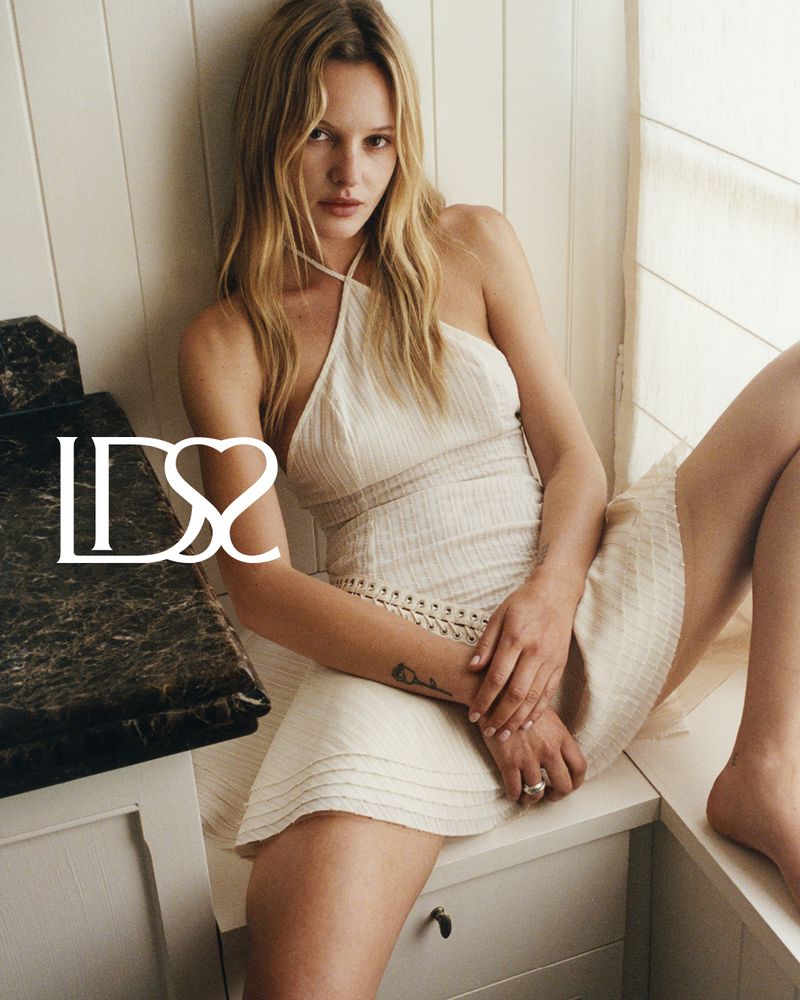 Model Paige Reifler wearing a white halter dress seated indoors against a panelled wall for Ludovic de Saint Sernin