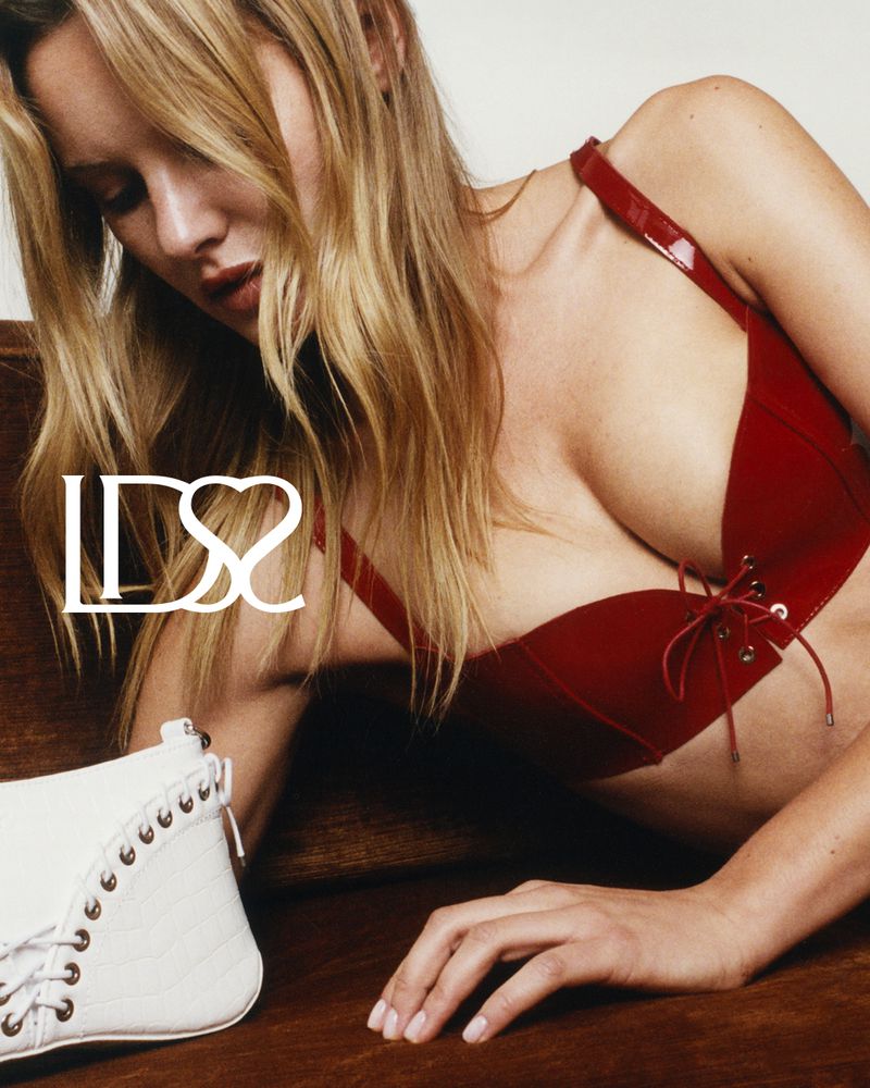 Blonde woman wearing a red bra top, laying on a wooden surface with a white shoe, indoor medium shot, fashion editorial style.