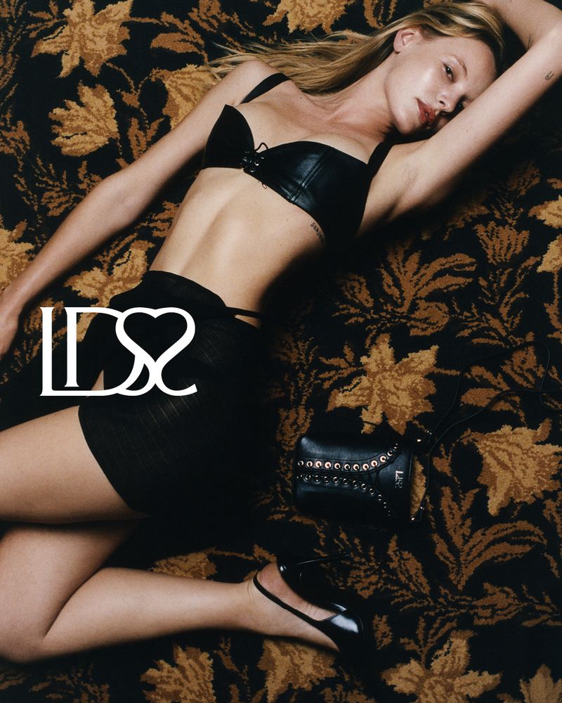 Blonde woman in black lingerie lies on a dark floral rug next to a black purse and shiny black heels, creating a fashion-forward scene.