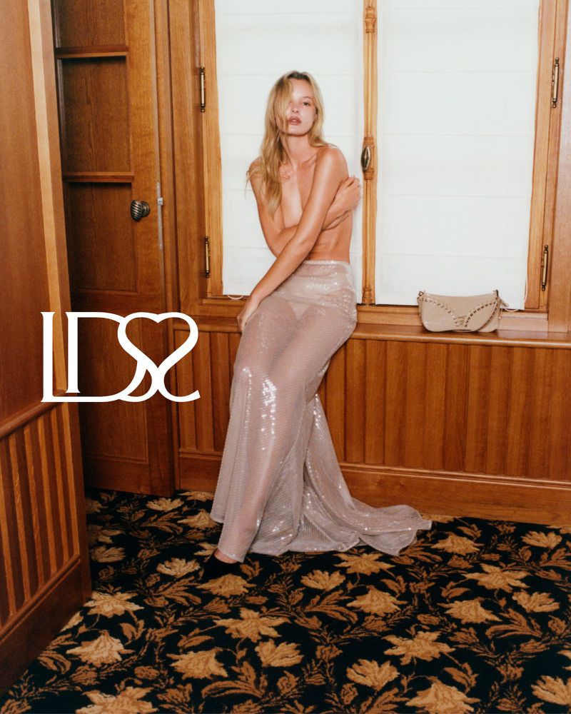 Fashion shot of a woman in a sequined skirt sitting near a window in a wood-paneled room, featuring a tan purse on the window sill.