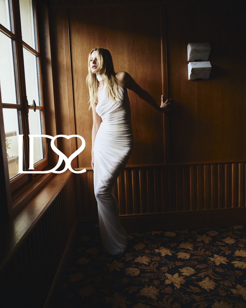 Paige Reifler in a white halter dress standing near a wooden wall and window for Ludovic de Saint Sernin S/S 25.