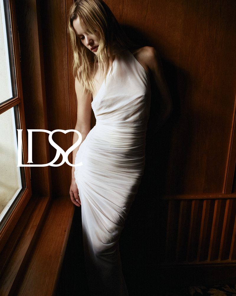 A fair-skinned woman in a white dress poses next to a window with a wood frame and paneling in an indoor setting.