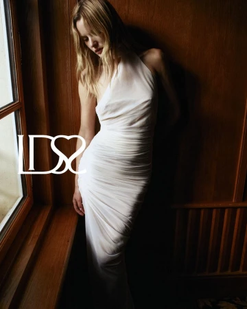 Model Paige Reifler wearing a white halter dress posing indoors against a dark wooden wall by a window.