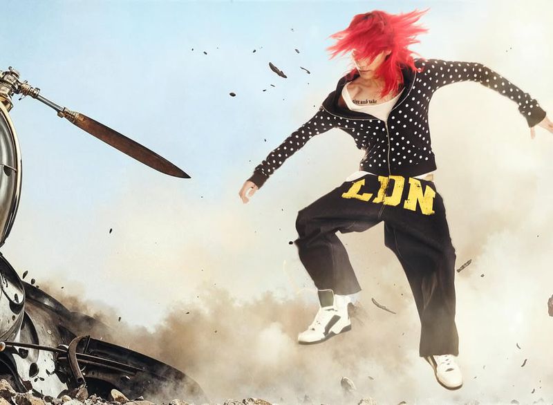 Male dancer with red hair in mid-air, wearing a polka dot hoodie and pants with yellow lettering, against a sky and debris background.