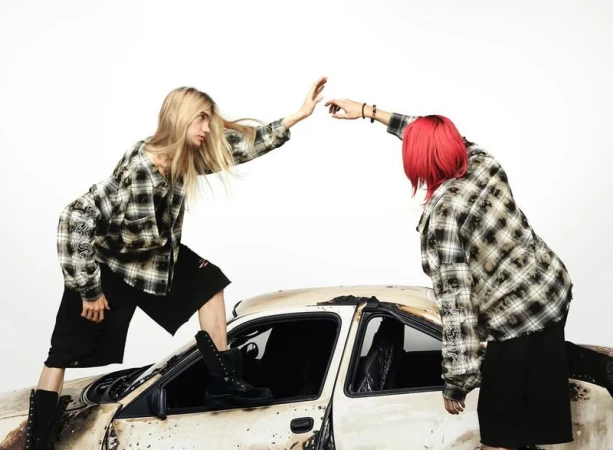 Two models in plaid shirts pose atop a distressed car in a studio, showcasing grunge fashion against a clean white background.