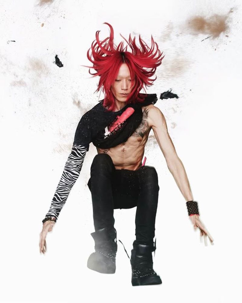 Man with red hair and expressive posing in a studio. He wears a black t-shirt, zebra sleeve, black pants, and boots against a white background.