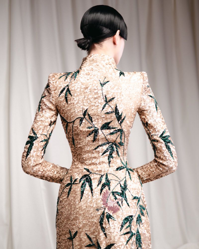Back view of a model wearing a shimmering gold, form-fitting gown with dark green floral vine embellishments, photographed against a neutral backdrop.