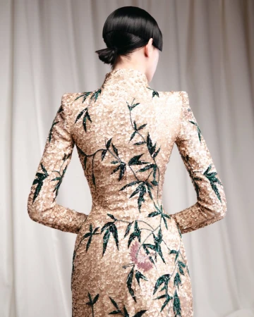 Model seen from behind wearing an embellished gold gown with green embroidered bamboo motifs against a cream curtain background