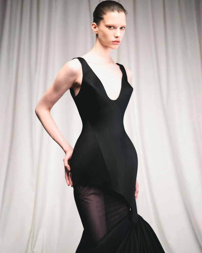 Elegant female model wearing a form-fitting black dress with sheer fabric detail, posing in a studio setting with soft, natural light.