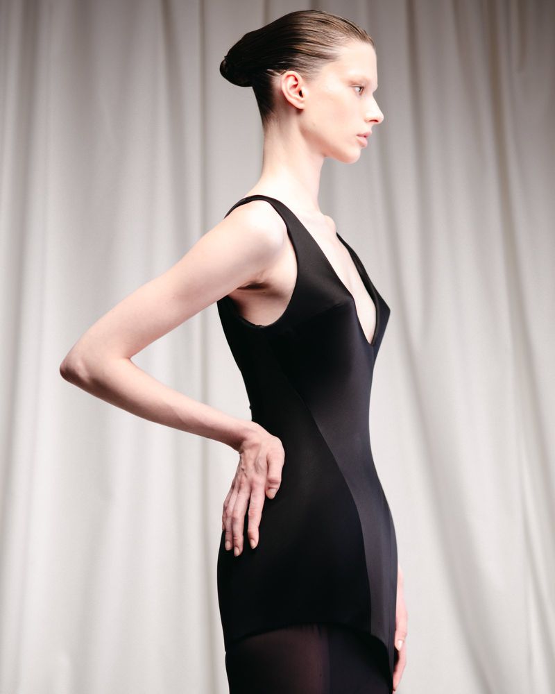 Woman in black sleeveless dress poses in front of neutral background. Fashion shot features side view of the adult female.
