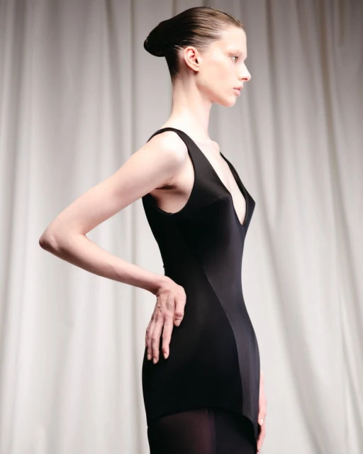 Woman in black sleeveless dress poses in front of neutral background. Fashion shot features side view of the adult female.