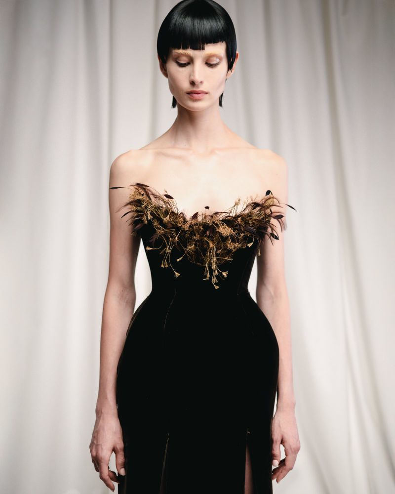 Elegant fashion portrait of a woman in a black strapless gown with feather details against a cream curtain backdrop in soft studio lighting.