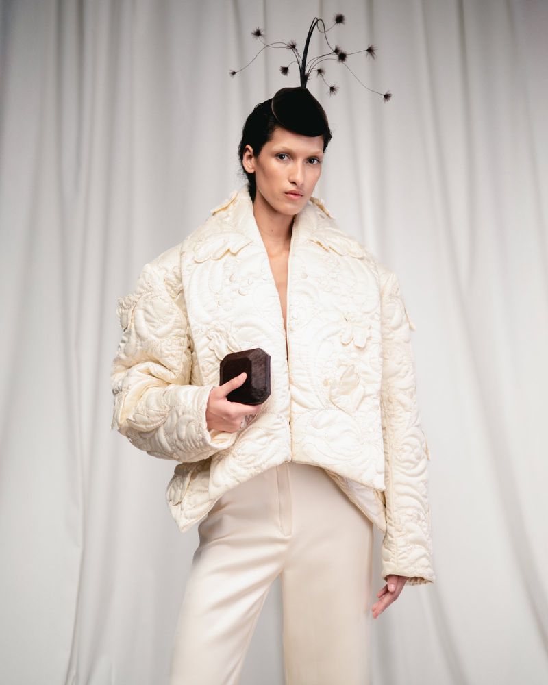 Adult female fashion model poses in a studio wearing a textured off-white jacket and trousers with a sculptural fascinator.