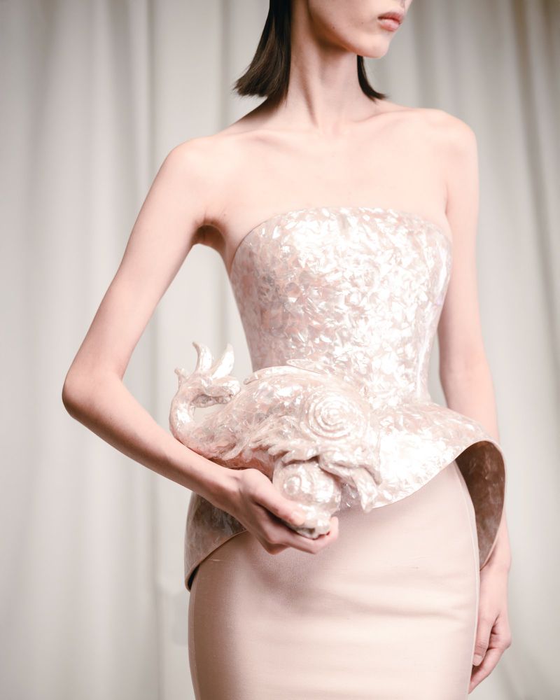 Woman in pale pink strapless dress with decorative sculpted elephant. Elegant couture fashion in a bright, clean studio setting.