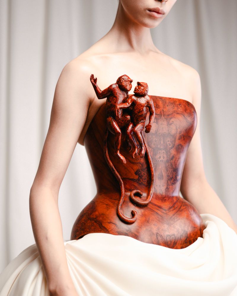 Fashionable woman in strapless dress with monkey sculpture on bodice against draped fabric backdrop, creating unique couture style.