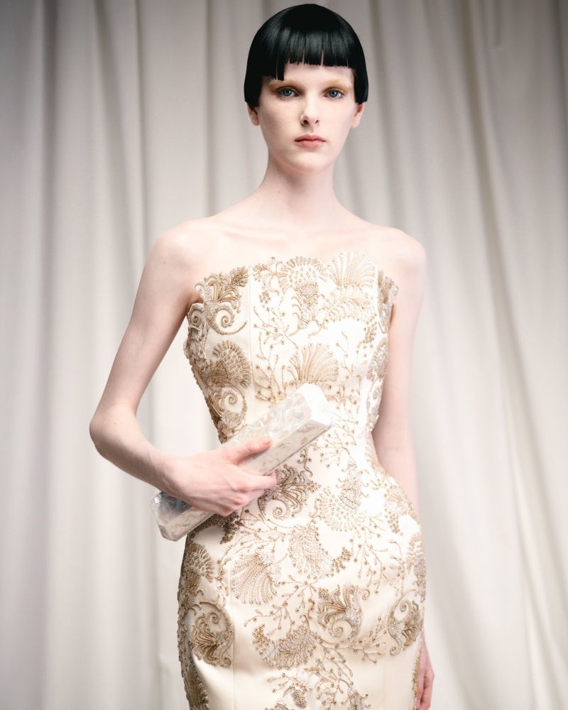 Woman in embellished strapless dress holding clutch against a white draped backdrop, studio shot for fashion editorial, neutral colors.