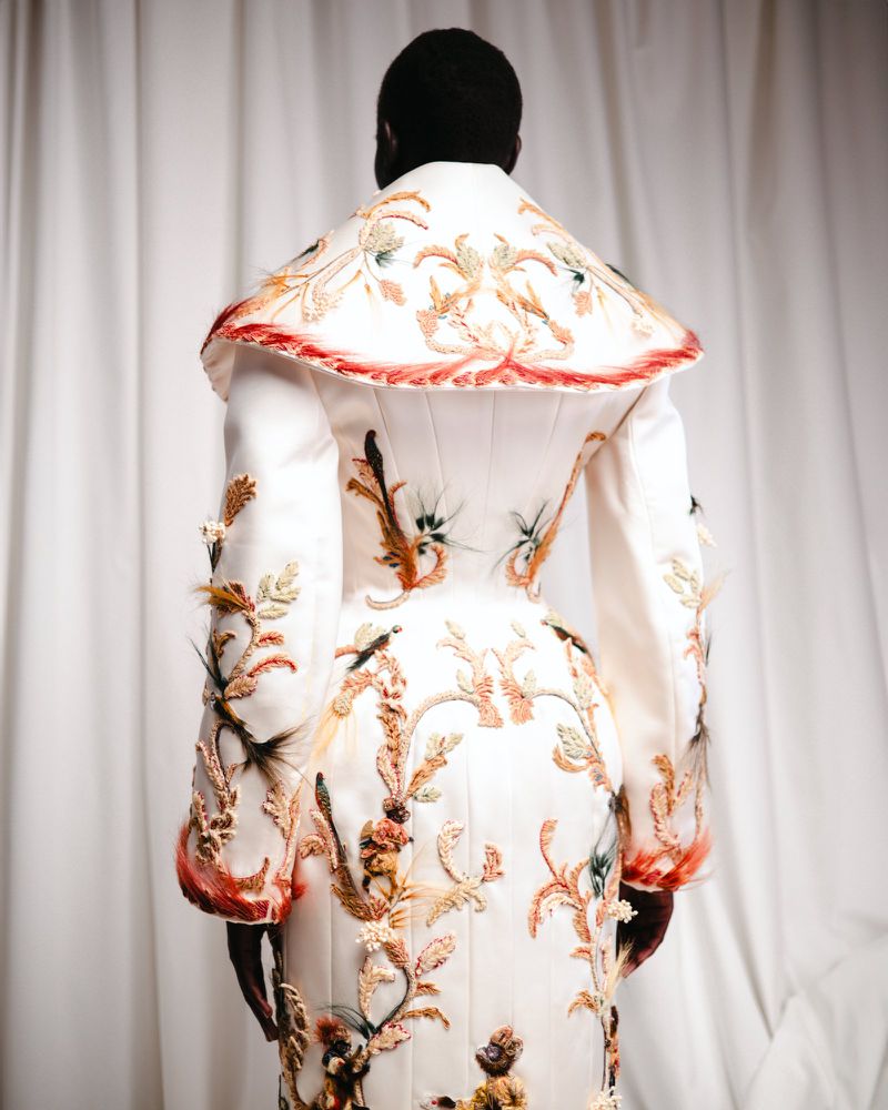 Model seen from behind wearing an ornate white couture coat with intricate embroidered and feathered patterns.