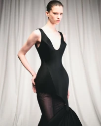 Model wearing a structured black evening dress posing against white pleated fabric curtains in a studio setting