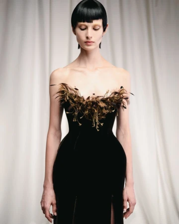 Model in a black strapless gown with feathered bodice and blunt bob hairstyle against a draped white curtain background.